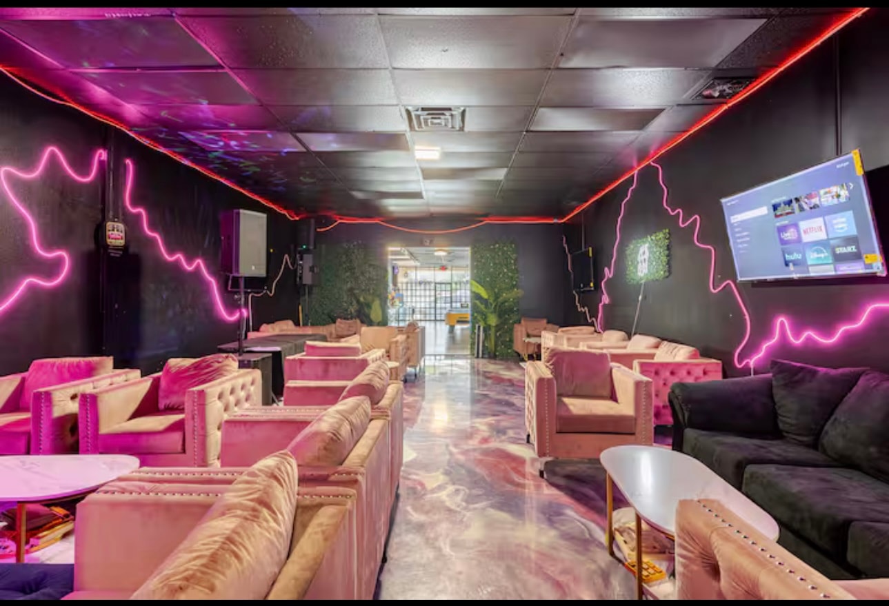 A lounge room lined with pink velvet seating, neon wall tubing and a large screen.