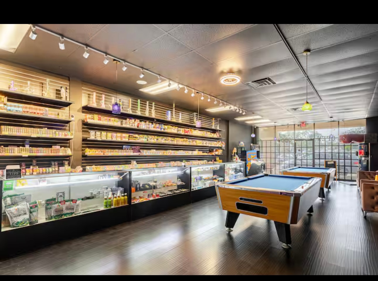 The retail floor: a long run of lit glass counter cases and a full wall of shelving stacked with vapes, e-liquid and hemp packaging, with the pool tables beyond.