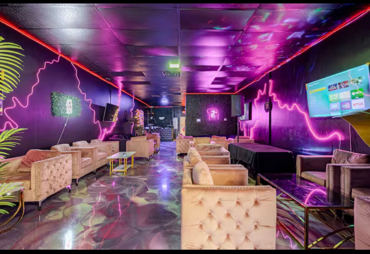 The lounge: tufted velvet armchairs on a poured metallic floor under pink and violet neon, with a wall-mounted TV.