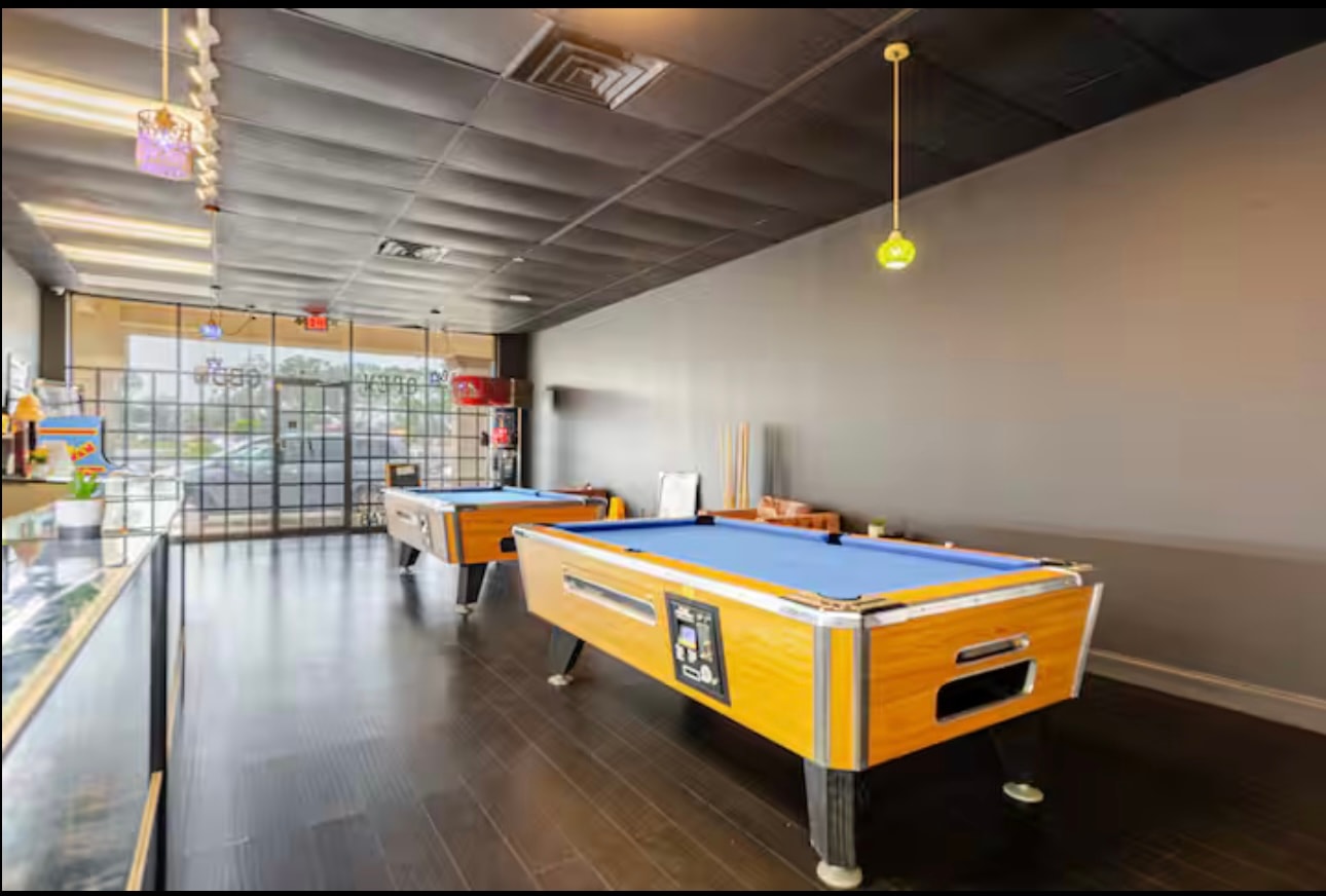 Two pool tables on the shop floor beside the counter cases, with the front windows behind them.